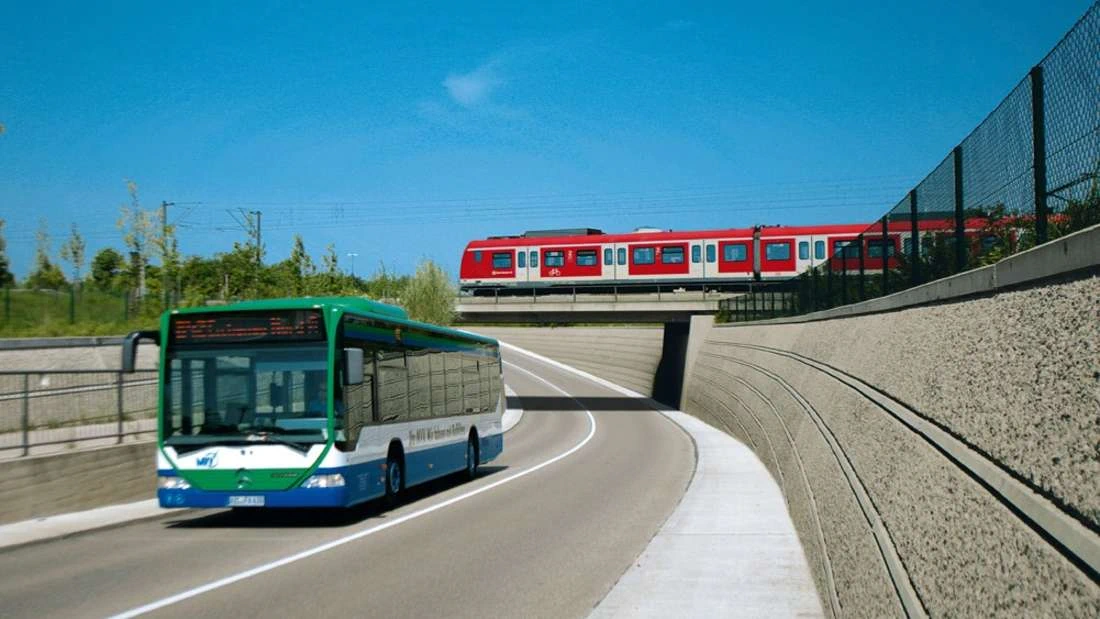 Foto eines Busses auf einer Stadtstraße. Im Hintergrund eine fahrende S-Bahn, die auf einer Brücke die Straße überquert.